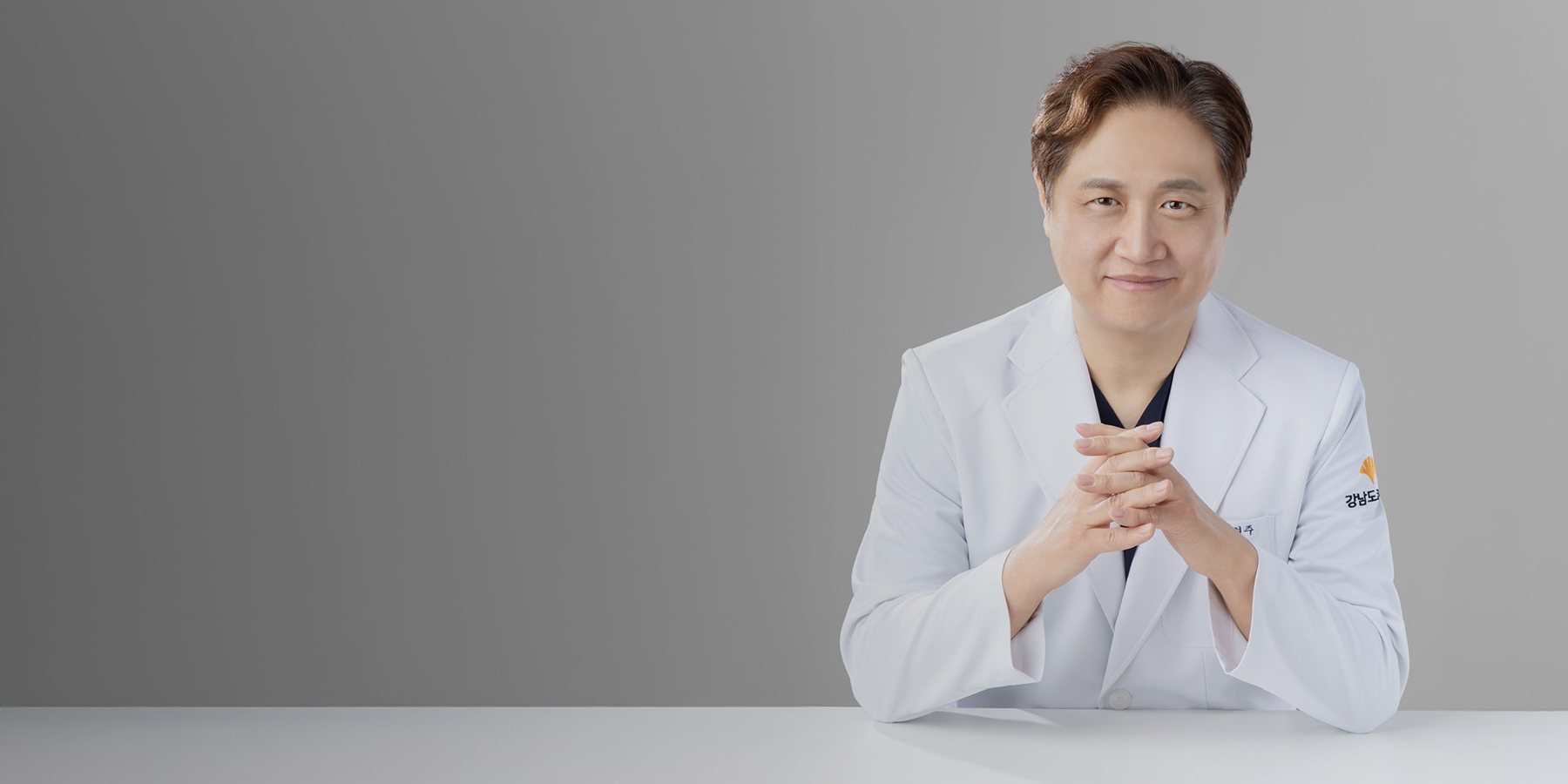 회색 배경, 흰 가운 차림으로 두 손을 깍지 낀 박형주 원장 상반신